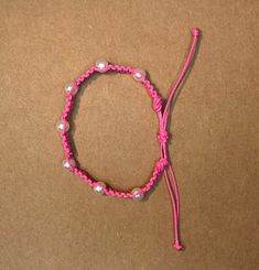 a pink string bracelet with white beads and a cord attached to the end, on a brown surface