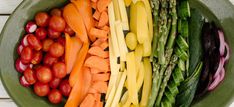 a bowl filled with different types of vegetables on top of a white wooden table next to tomatoes and asparagus