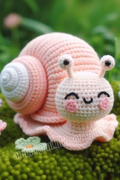 a small crocheted snail laying on top of green moss covered in tiny flowers