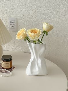 three yellow roses in a white vase next to a candle and a necklace on a table