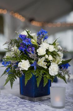 a blue vase with white flowers and greenery sits on a table next to a candle