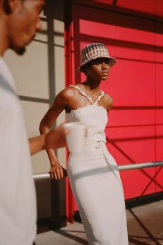 a woman in a white dress and hat standing next to a red wall with a man looking at her