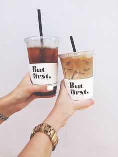 two people holding up cups with drinks in them that say but first, but first