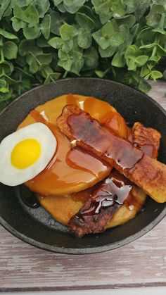 an egg is on top of pancakes with syrup and bacon in a black bowl next to some green leaves