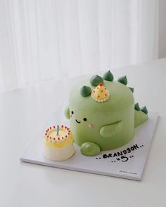 a green dinosaur cake sitting on top of a table next to a small cupcake