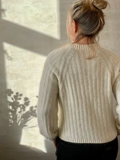 a woman standing in front of a window with her back turned to the camera, wearing a white sweater and black pants