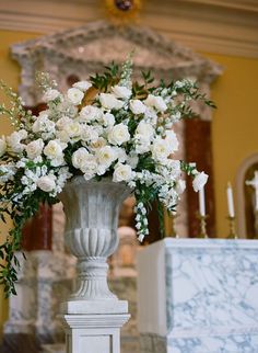 white flowers are in a large vase on a pedestal at the end of a church aisle