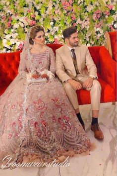 the bride and groom are sitting on a red couch in front of a floral wall