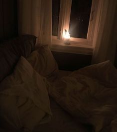 an unmade bed in front of a window with a lit candle on the windowsill