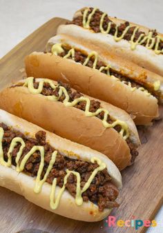 three chili cheese dogs with mustard drizzled over them on a cutting board