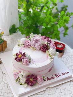 a white cake with pink and purple flowers sitting on top of a table next to a book