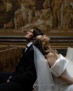 a bride and groom sitting on a bench in front of a wall with paintings behind them