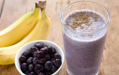 a glass of blueberry smoothie next to two bananas on a wooden table with other ingredients