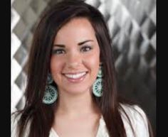 a woman with long brown hair and green earrings smiles at the camera while wearing a white shirt