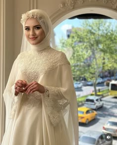 a woman in a white dress and veil standing next to a window with cars on the street