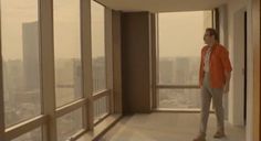 a man standing in an empty room looking out at the city from his apartment window