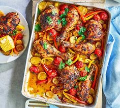 a pan filled with chicken and vegetables next to a plate of corn on the cob