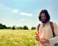 a man with long hair and beard holding a flower in his hand while standing in a field