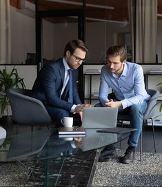 two men in suits sitting at a glass table looking at a laptop computer, both on their knees