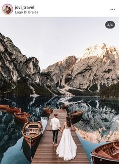 a bride and groom walking on a dock next to boats in the water with mountains in the background