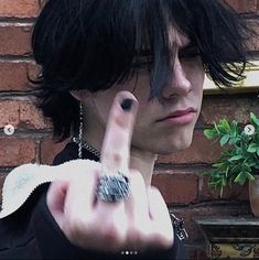 a man with black hair and piercings making the peace sign in front of a brick wall