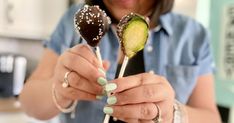 a woman holding two chocolate covered spoons with cucumbers on them