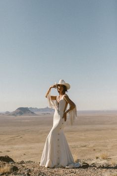 a woman in a white dress and hat standing on top of a dirt hill with mountains in the background