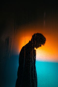 a man standing in front of a window covered in drops of water at sunset with his head down