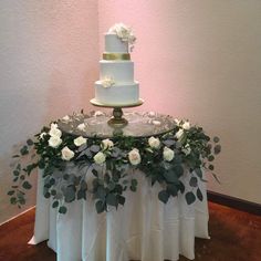 a three tiered cake with white flowers and greenery on the top is sitting on a table