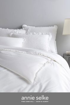 white bedding with ruffled trim and pillows in a bedroom setting on a grey wall