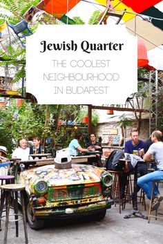 people sitting at tables in front of an old car with graffiti on it and the words jewish quarter