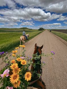 two people riding horses on a dirt road with flowers in the foreground and an open field behind them