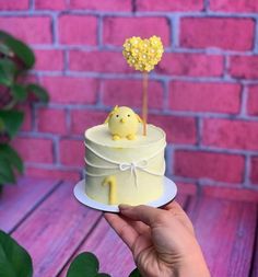 a hand holding a small cake with a yellow bird on top and a pink brick wall in the background