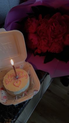 a birthday cake in a box with a candle on it next to some pink flowers