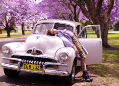 a woman laying on the hood of an old car in front of trees with purple flowers