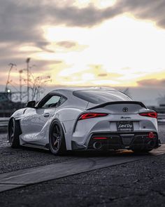 the rear end of a white sports car parked on top of a parking lot at sunset