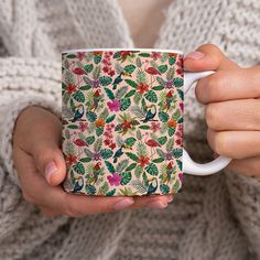 a woman is holding a coffee mug with green leaves and flowers on the outside, while wearing a white sweater