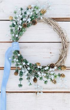 a wreath with pine cones, white flowers and blue ribbon hanging on a wooden wall