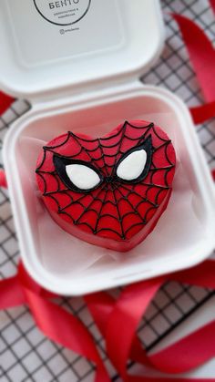 a spiderman cookie in a white container with red ribbon on the table next to it