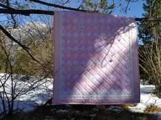 a pink and white quilt hanging from a tree in the snow with trees behind it