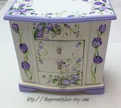 a white and purple flowered cabinet with drawers on it's sides, sitting on a table