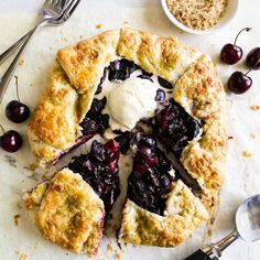 a pie with cherries and ice cream on top next to spoons, fork and bowl