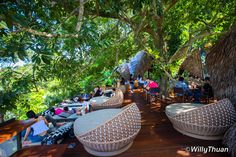 people are sitting at tables under the shade of some trees and grass umbrellas on a wooden deck