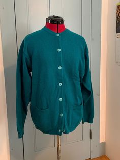 "This classic pocket cardigan by McKenzy is made of green-teal virgin wool. It has two pockets on the front and closes with 7 pearlescent plastic buttons. It has a spare button sewn inside. It measures approximately 45\" unstretched at the chest. The center back length is approximately 27\", the sleeves are approximately 23\" and the neck to shoulder is approximately 7\". It is in good condition. There is some discoloration on the underside of the sleeves. I am not sure if it is a stain. (It almost looks like dye settling from being washed.)" Classic Green Wool Cardigan, Green Button-up Cardigan With Pockets, Vintage Green Cardigan With Button Closure, Green Crew Neck Sweater With Pockets, Green Button-up Sweater With Button Closure, Green Long Sleeve Sweater With Pockets, Pocket Cardigan, Sewing A Button, Wool Sweater