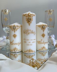 three white candles with gold embellishments are on a table next to wine glasses