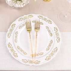 two forks are placed on a white plate with gold trim around the edges, along with an empty glass vase filled with flowers