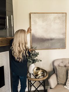 a woman standing in front of a painting next to a chair and table with a potted plant on it