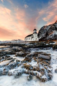 a white church sitting on top of a snow covered mountain next to the ocean at sunset