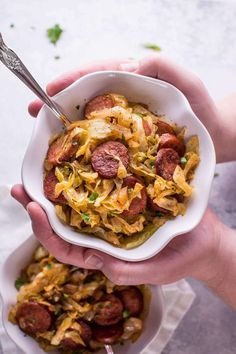 two hands holding a bowl of food with sausage, cabbage and noodles on the side