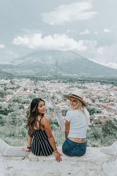 two women sitting on top of a stone wall looking at the city in the distance